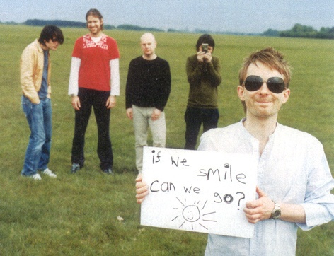The five members of Radiohead. Four of them are in the background; in the lower right corner you have a man holding a sign that reads ‘if we smile, can we go?’.